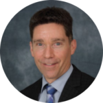 Sam Demma A man in a suit and tie poses for a professional headshot against a dark, blurred background, representing the polished image of Elite Speaker Coaching with Sam Demma. Elite Speaker Coaching