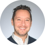 Sam Demma Man with short curly hair and light facial hair, wearing a white shirt and dark jacket, smiling at the camera against a light grey background—perfectly capturing Sam Demma’s approachable style as part of Elite Speaker Coaching. Elite Speaker Coaching