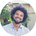 Sam Demma A man with curly hair and a beard, wearing a white shirt with a blue pattern, smiles outdoors with greenery and a building in the background—perfectly capturing the approachable energy of Sam Demma from Elite Speaker Coaching. Elite Speaker Coaching
